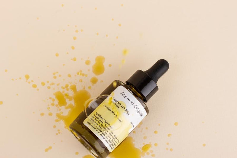 Apprenti organik natural facial oil laid across peach background with facial oil splatter across bottle and background