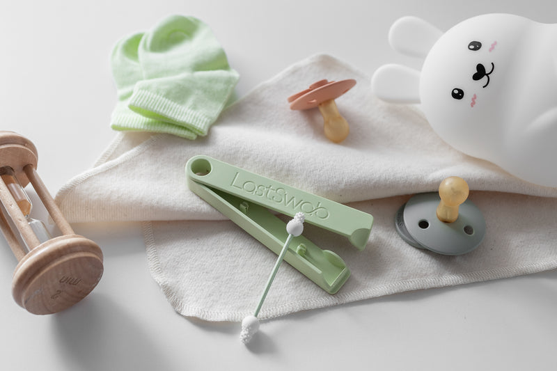 Reusable green baby ear swab next to baby toys, soother and cloth