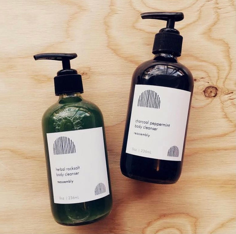 reassembly botanical green herbal rock salt cleanser and black charcoal peppermint cleaner laid on wooden background