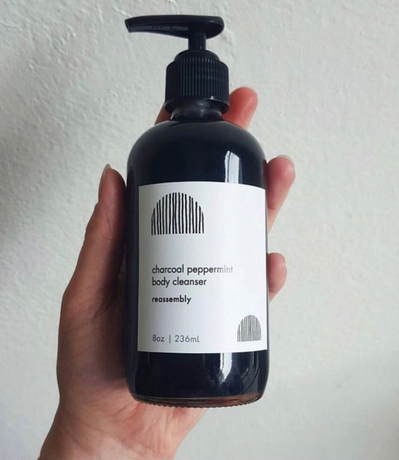 women's hand holding glass pump bottle of reassembly botanical charcoal peppermint body cleanser