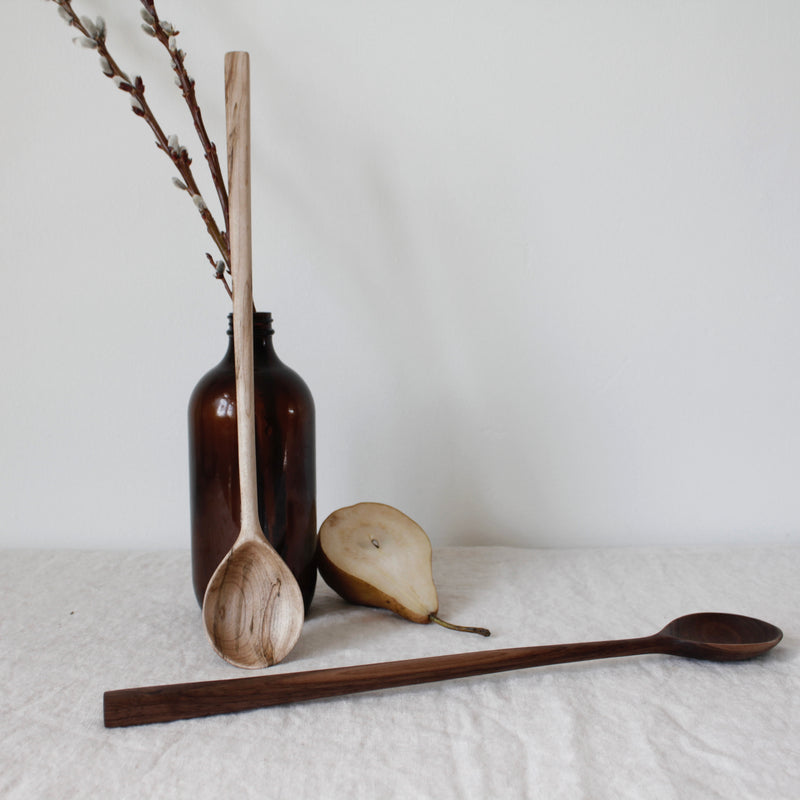 the woodsmans daughter walnut and maple hand carved hardwood stirring spoons laid on table and against amber bottle